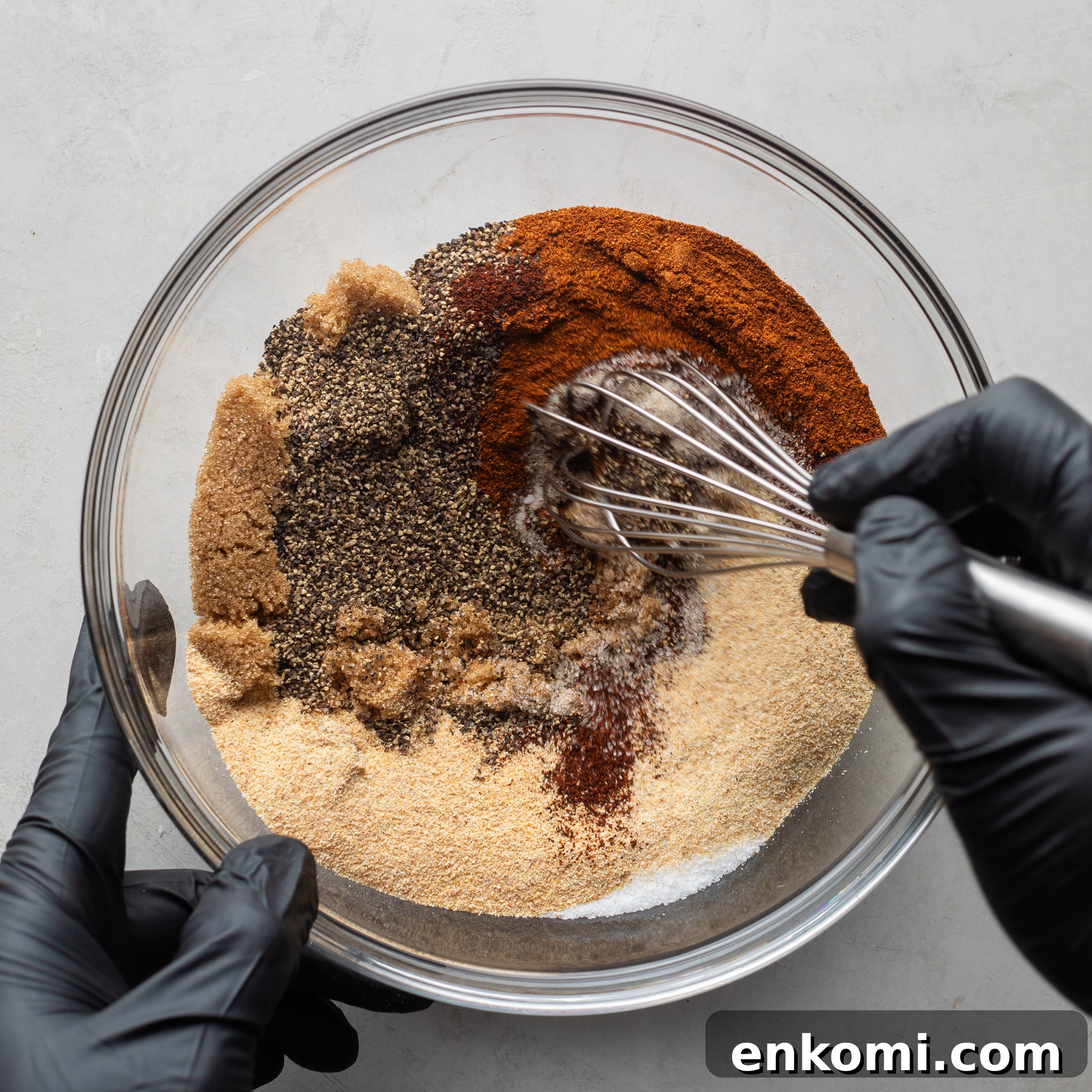 Homemade BBQ Rub Recipe for Tender, Flavorful Grilling 5 A hand wearing black latex gloves mixing with a whisk the BBQ seasoning ingredients in a large glass bowl.