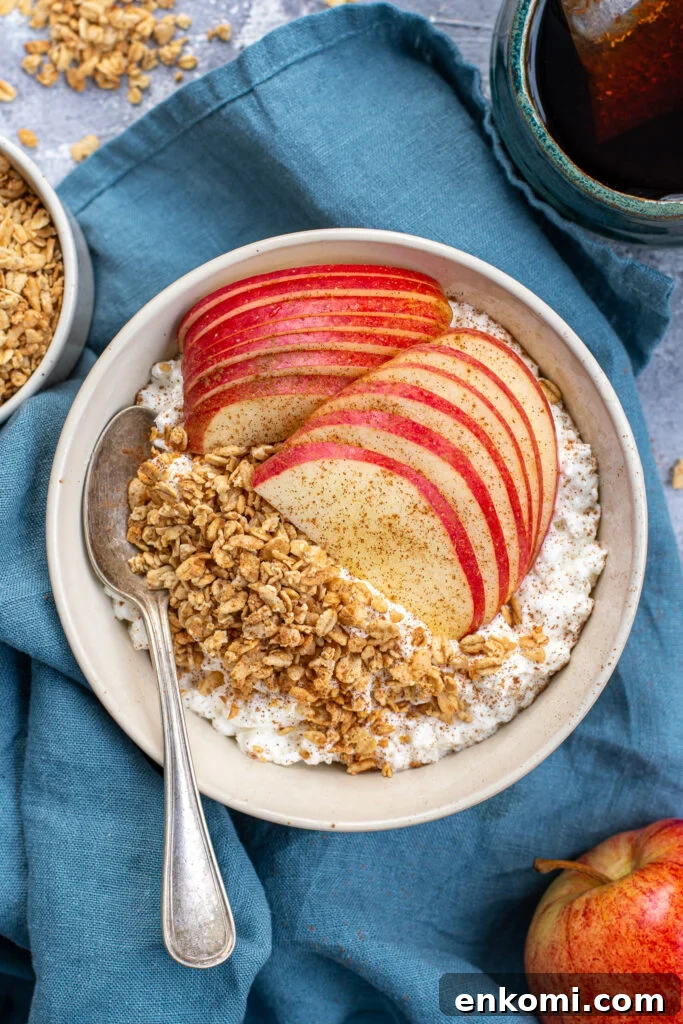 Cottage Cheese & Apple Breakfast Bowls with Granola and Honey 2 A white bowl on a blue towel filled with cottage cheese and apples with granola and ground cinnamon with a spoon ready to eat.