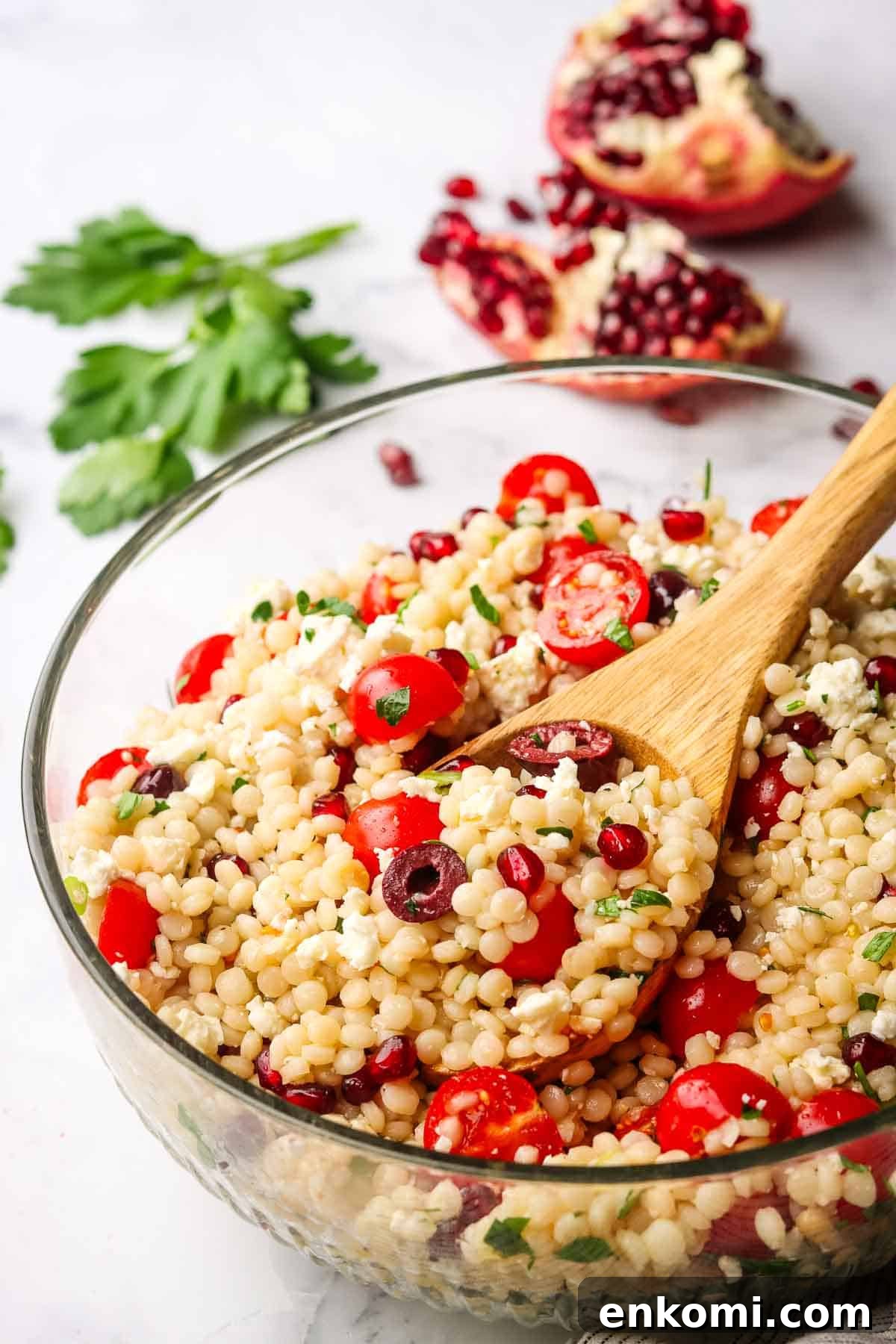 Mediterranean Pearl Couscous Salad with Fresh Herbs & Feta 11 A large bowl of Israeli couscous salad with a wooden spoon in it. Pomegranate and parsley in the background.