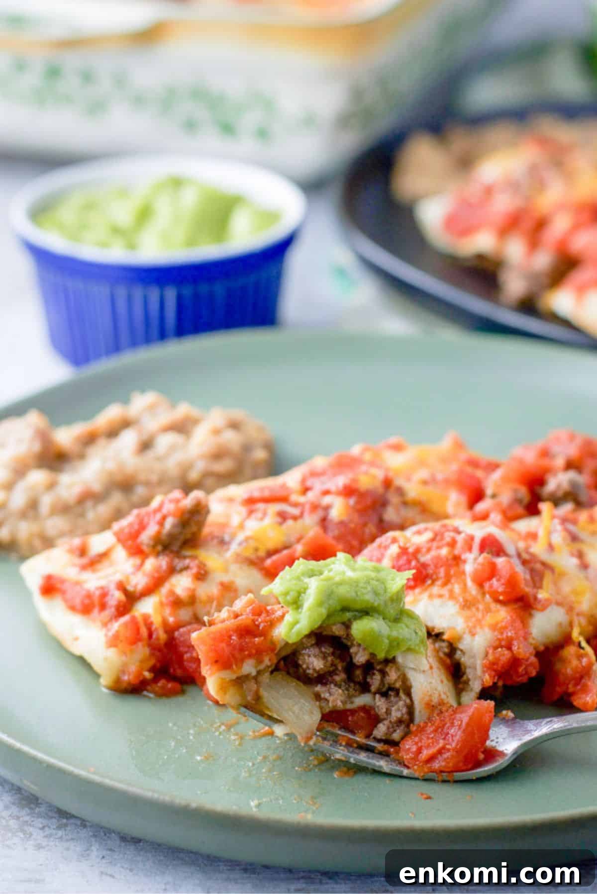A fork with a bite of enchilada resting on a plate with two enchiladas and beans. There is guac and more enchiladas in the background