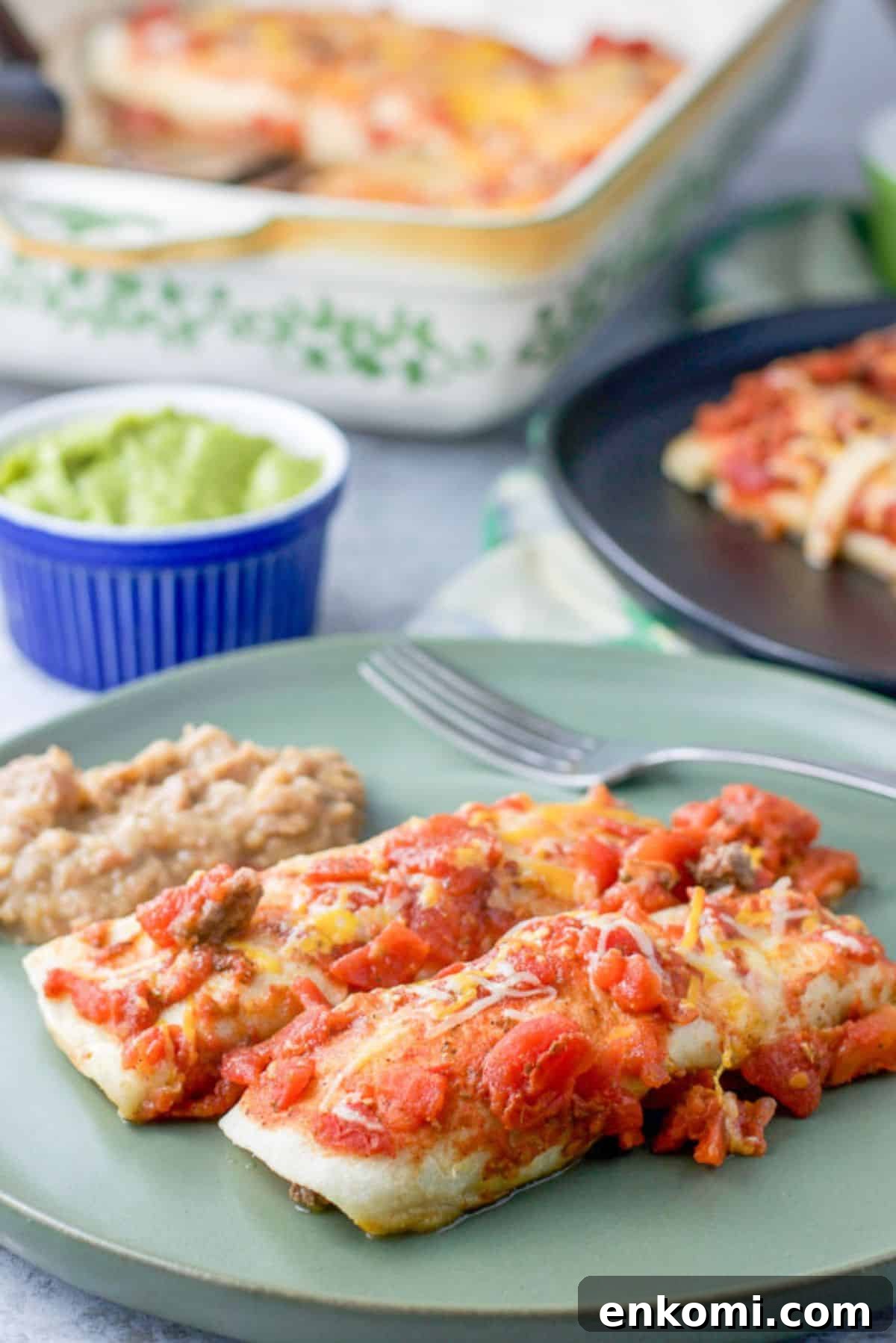 Two enchiladas with a red sauce on a plate with some beans. There is some guacamole in the background along with another plate of enchiladas
