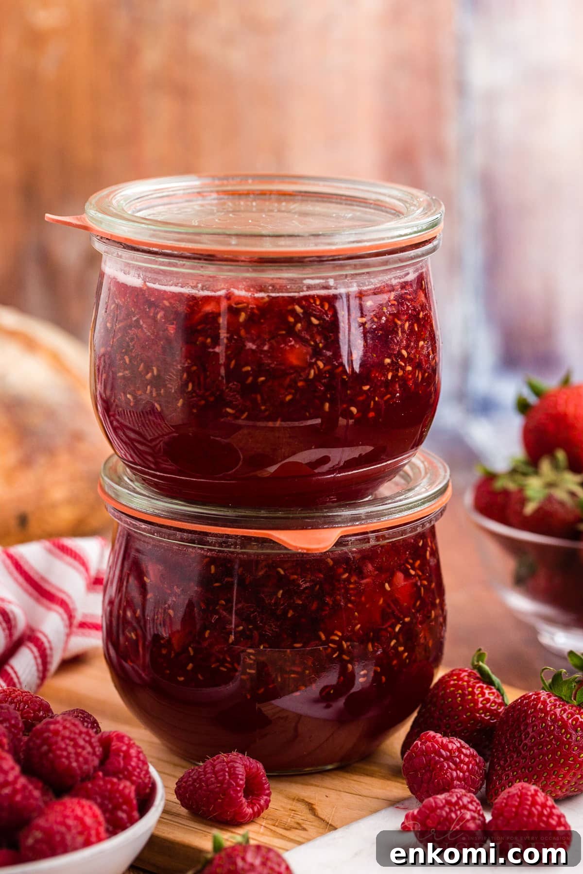 Two stacked glass jars filled with slow cooker strawberry and raspberry jam, on top a wooden board, bowl of fresh raspberries and loose raspberries and strawberries on a wooden surface, red and white striped linen cloth