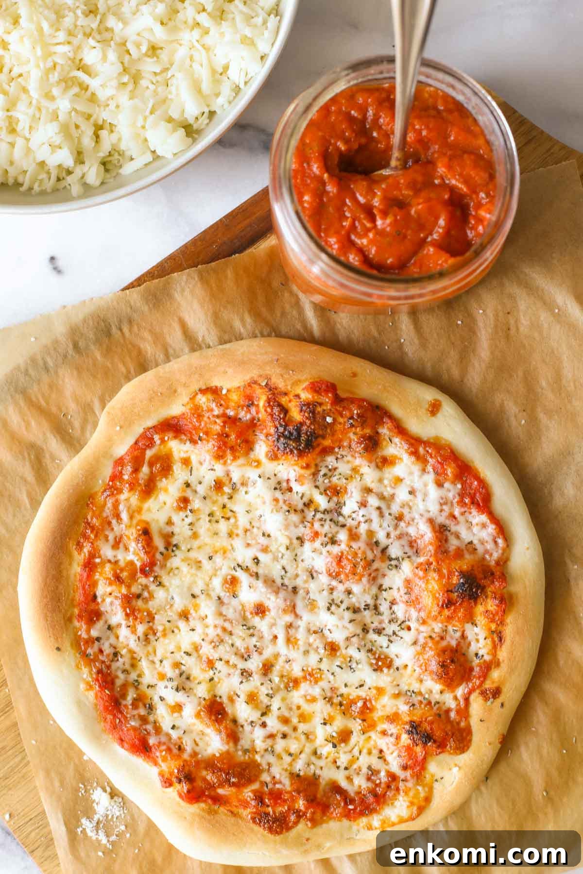 Roasted Tomato Pizza Sauce Recipe for Rich, Smoky Flavor 2 Overhead shot of a homemade cheese pizza on parchment paper, sitting on a wood cutting board with a jar of Roasted Tomato Pizza Sauce, and a bowl of shredded mozzarella cheese next to the cutting board.