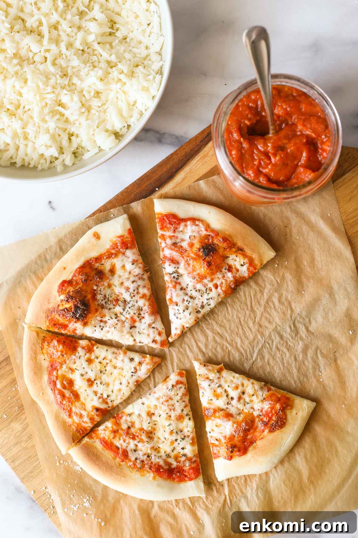 Roasted Tomato Pizza Sauce Recipe for Rich, Smoky Flavor 11 Overhead shot of slices of homemade cheese pizza on parchment paper, sitting on a wood cutting board with a jar of Roasted Tomato Pizza Sauce, and a bowl of shredded mozzarella cheese next to the cutting board.