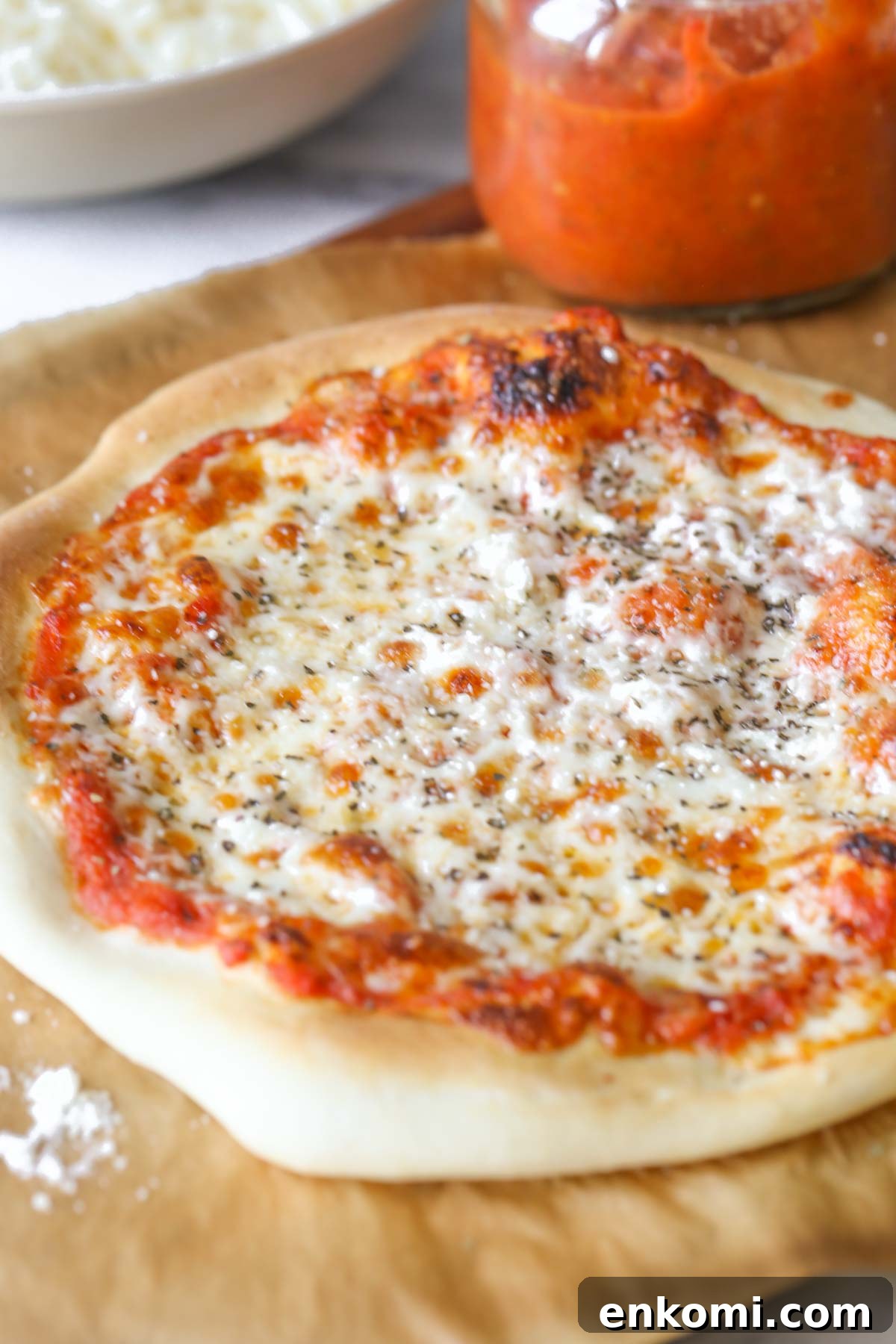 Roasted Tomato Pizza Sauce Recipe for Rich, Smoky Flavor 10 Close up shot of a homemade cheese pizza on parchment paper, sitting on a wood cutting board with a jar of Roasted Tomato Pizza Sauce.