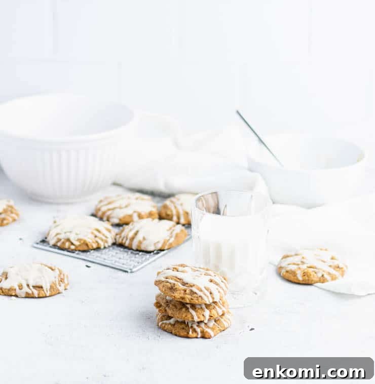 Iced pumpkin oatmeal cookies stacked in front of a cooling rack of cookies, and white bowls.
