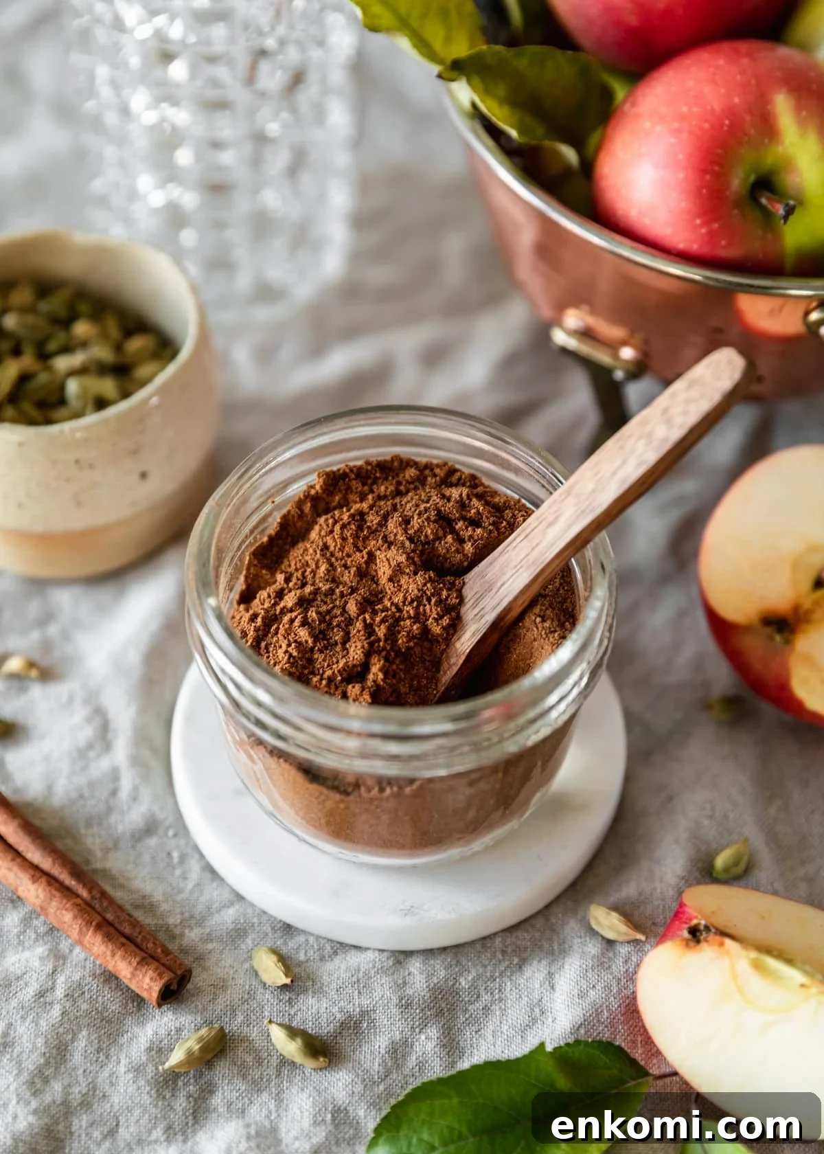 A side image of a jar of homemade apple pie spice on a beige linen next to a copper colander, sliced apple, cream bowl of cardamom pods, and a cinnamon stick.