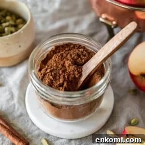 A side image of a jar of apple pie spice on a beige linen next to a white bowl of cardamom pods, a cinnamon stick, and sliced apples.