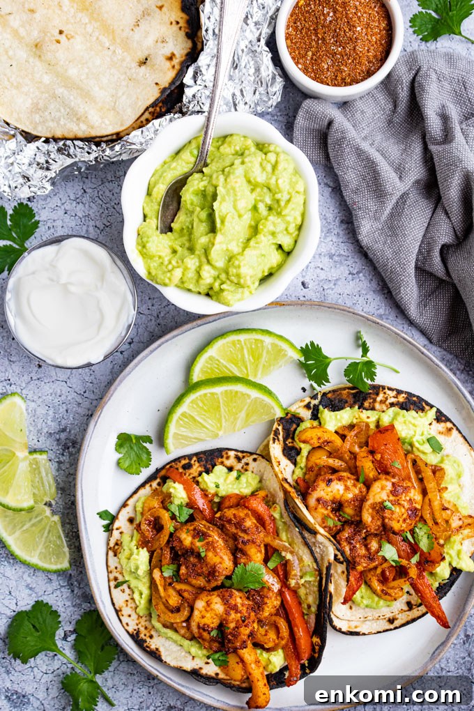Two shrimp fajitas on a gray plate with limes. A bowl of guacamole, sour cream, and toasted tortilla shells are in the background.