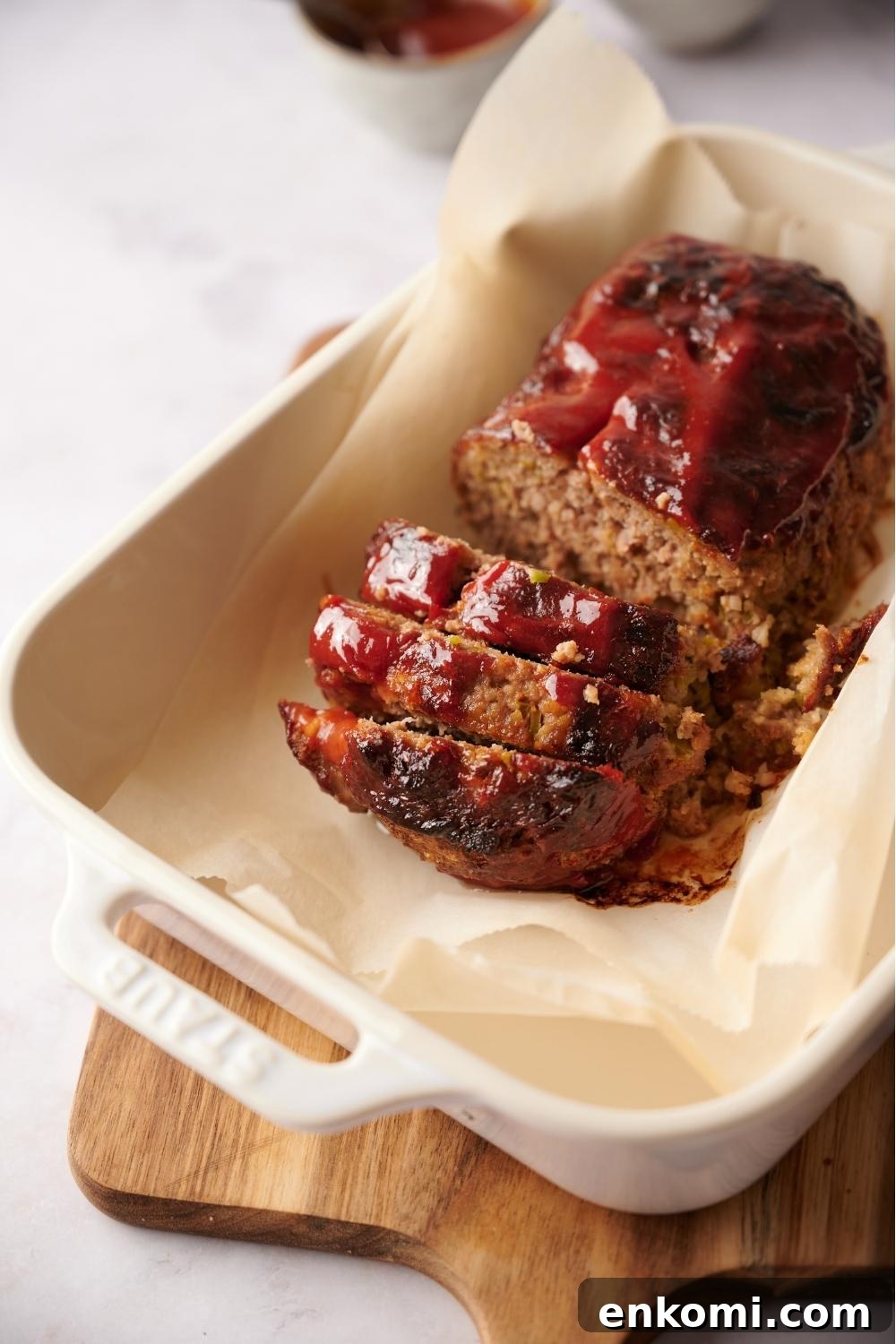 Southern meatloaf on parchment paper in a white baking dish. The dish is resting on a wood cutting board. The meatloaf has been slice to serve and layers are stacked on top of one another.