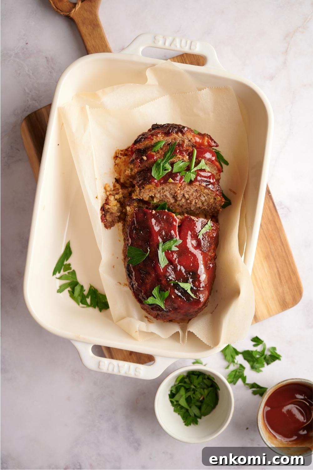 Southern meatloaf on parchment paper in a white baking dish. The dish is resting on a wood cutting board. Half of the meatloaf has been sliced to serve and the meatloaf is garnished with fresh parsley. Half-empty bowls of parsley and ketchup lay next to the meatloaf.