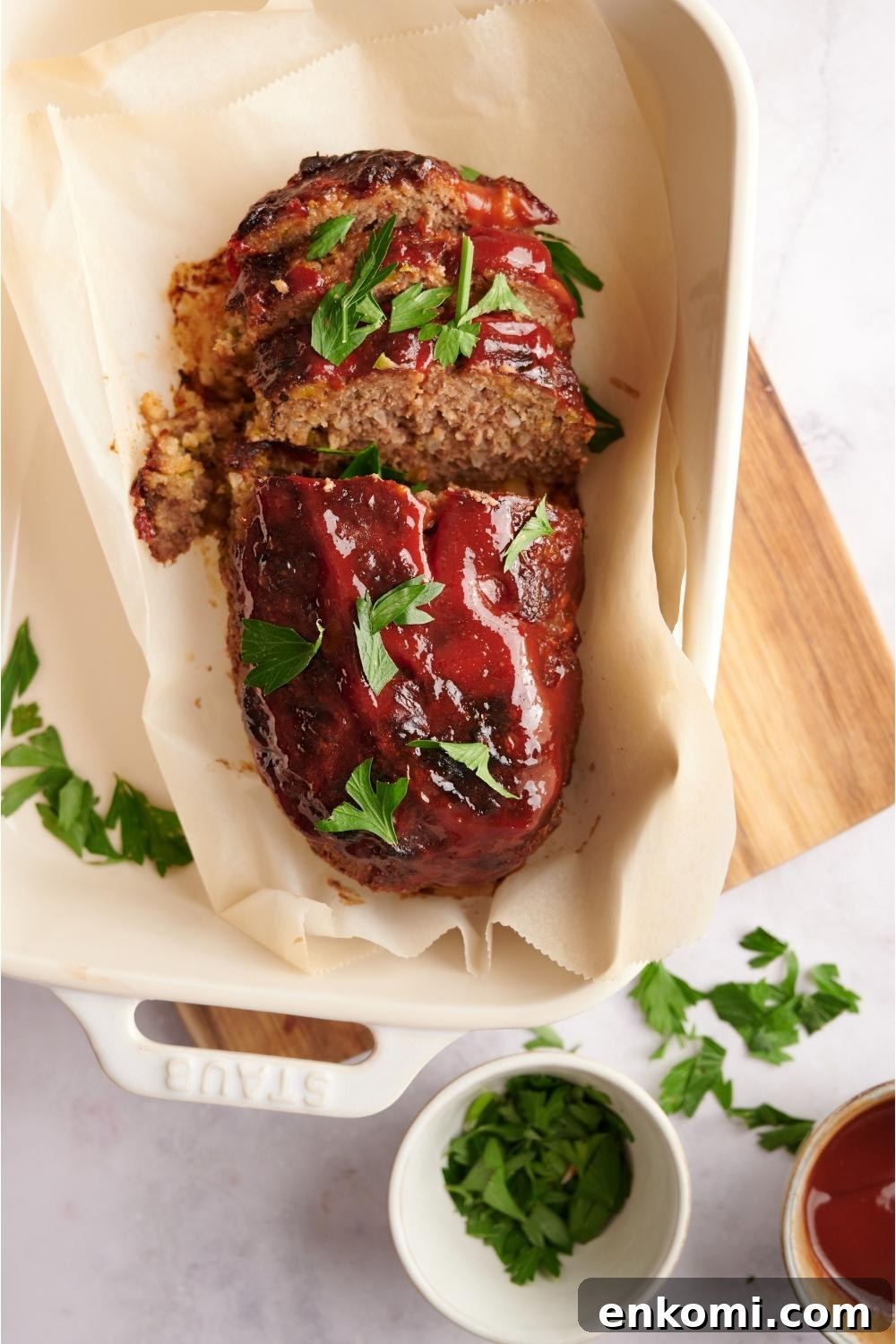Southern meatloaf on parchment paper in a white baking dish. The dish is resting on a wood cutting board. Half of the meatloaf has been sliced to serve and the meatloaf is garnished with fresh parsley. A half-empty bowl of parsley is next to the meatloaf.