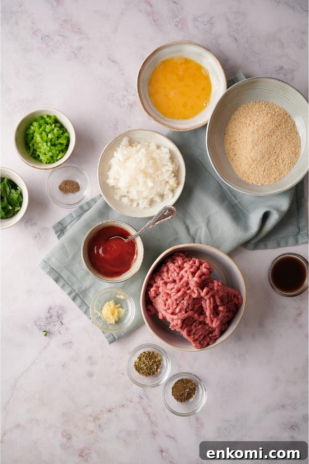 An assortment of ingredients including bowls of bread crumbs, egg, chopped onion, raw ground beef, ketchup, and seasonings. Most of the bowls are on a blue dish towel and everything is on a grey counter.