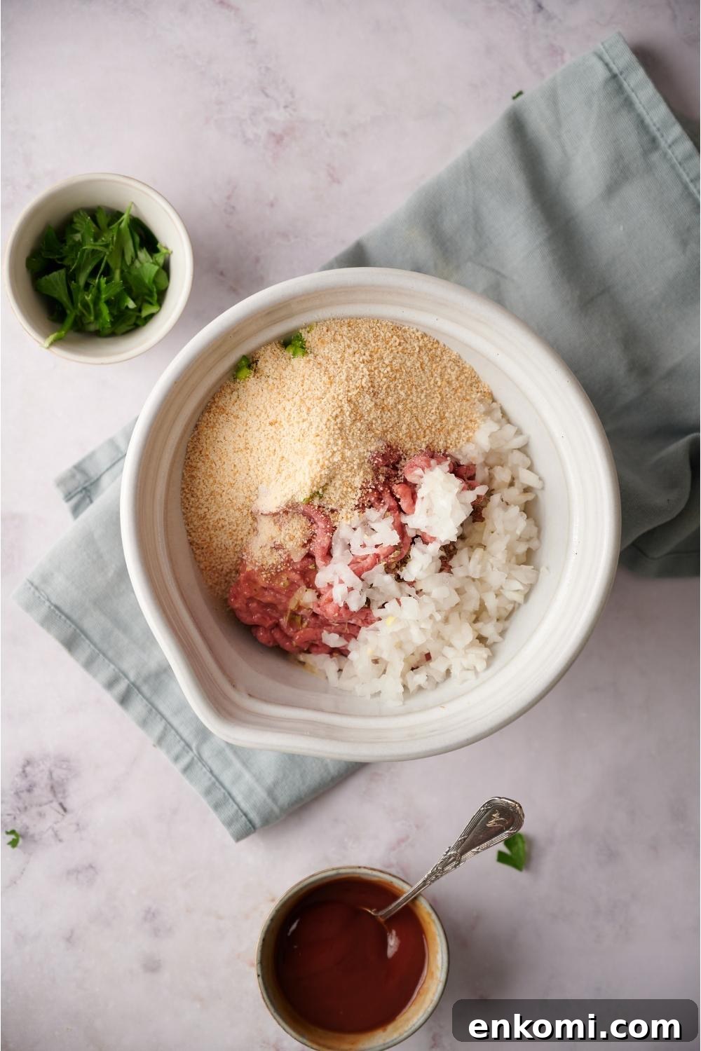 A large white mixing bowl with all of the raw ingredients to make southern meatloaf in the bowl. The ingredients have not yet been mixed together. The bowl is on a blue dish towel on a grey counter and bowls of ketchup and parsley lay next to the bowl. There is a spon in the bowl of ketchup.
