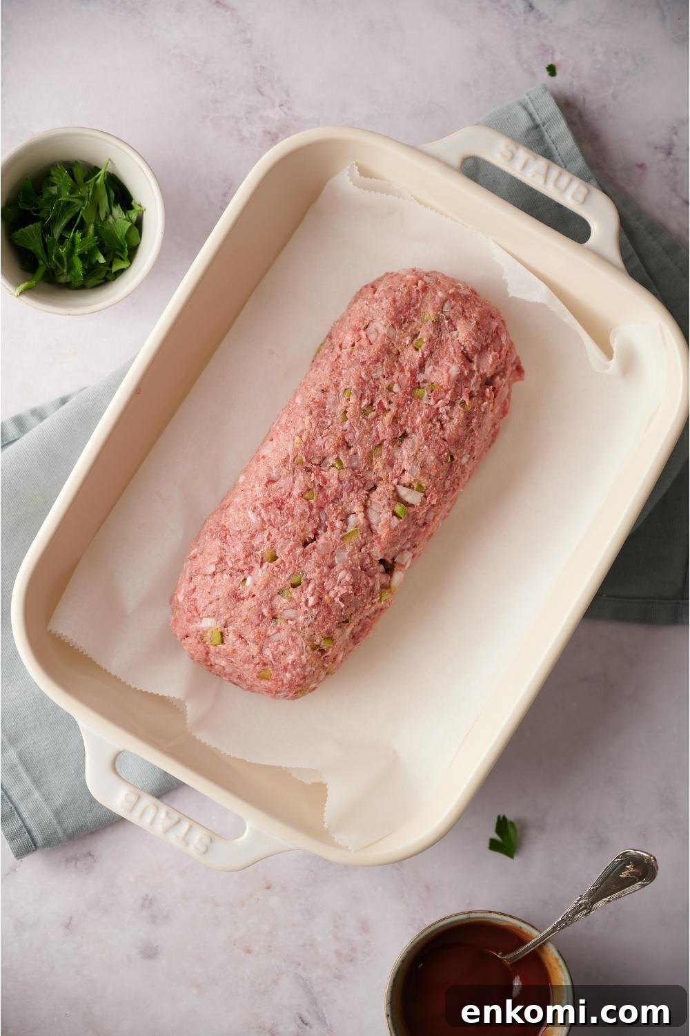 An uncooked loaf of southern meatloaf on parchment paper in a white baking dish. The dish is on a blue dish towel on a grey counter. Bowls of parsley and ketchup lay next to the meatloaf with a spoon in the ketchup bowl.