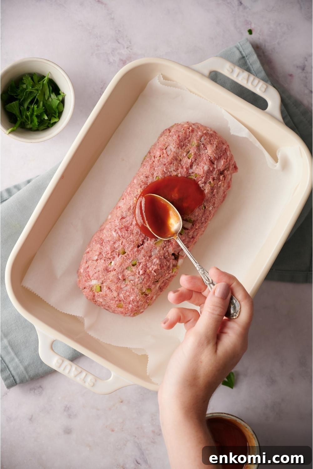 An uncooked loaf of southern meatloaf on parchment paper in a white baking dish. A hand is spooning ketchup on the loaf. The dish is on a blue dish towel on a grey counter and a bowl of parsley is next to the dish.