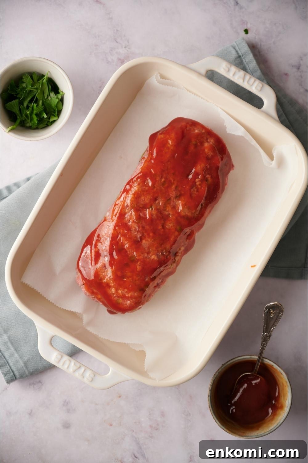 An uncooked loaf of southern meatloaf on parchment paper in a white baking dish with ketchup covering the meatloaf. The dish is on a blue dish towel on a grey counter and bowls of parsley and ketchup lay next to the dish. There is a spoon in the ketchup bowl.