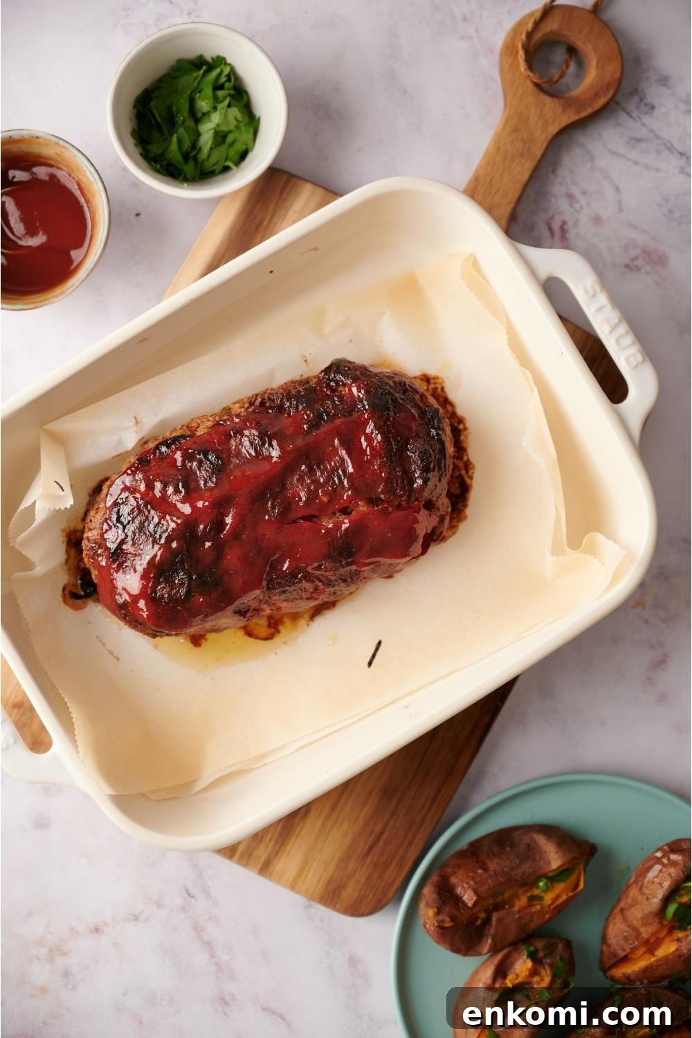 Cooked southern meatloaf on parchment paper in a white baking dish on a wooden cutting board. Bowls of ketchup and parsley are next to the dish and a plate of sweet potatoes are to the right of the cutting board.