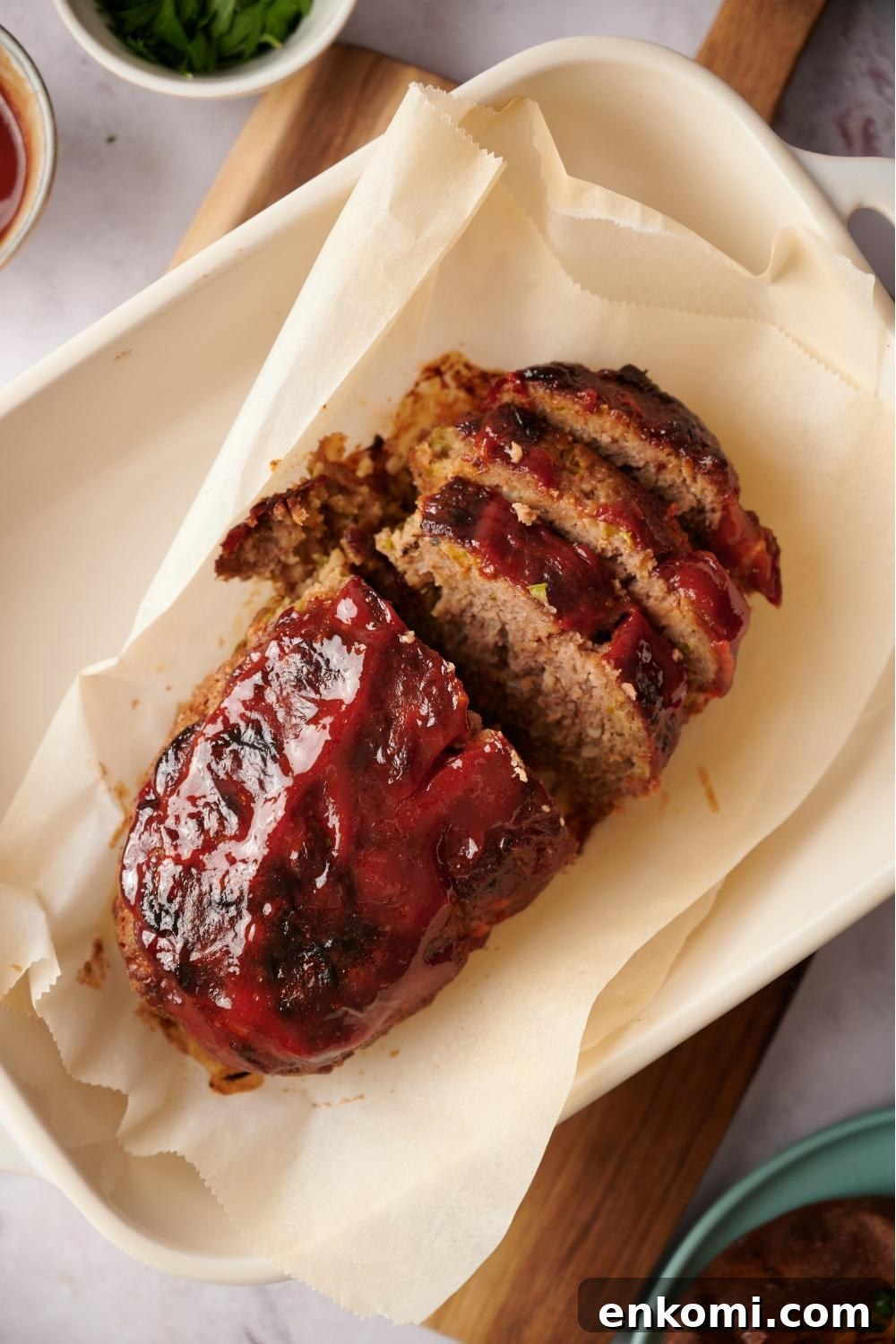 Southern meatloaf on parchment paper in a white baking dish. The dish is resting on a wood cutting board. Half of the meatloaf has been sliced to serve.