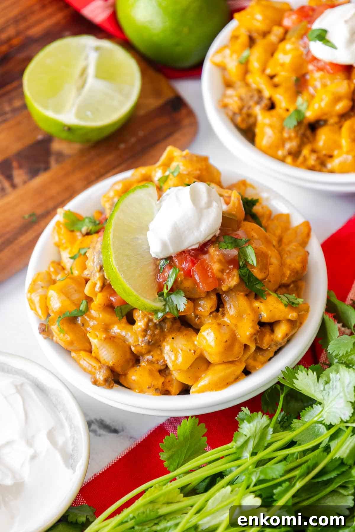 A close-up bowl of taco mac and cheese topped with salsa, sour cream, fresh cilantro, and a lime wedge. The creamy cheese shells are mixed with seasoned ground beef.