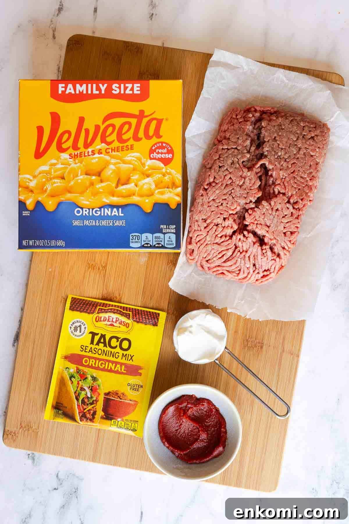 Ingredients for taco mac and cheese laid out on a wooden board, including a box of Velveeta shells and cheese, raw ground beef, taco seasoning packet, tomato paste, and sour cream.