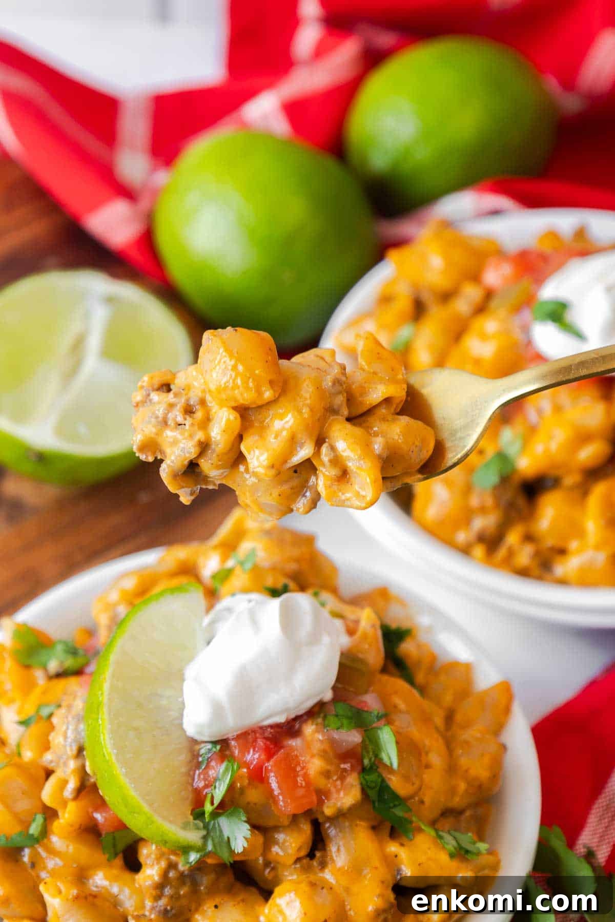 A forkful of taco mac and cheese held above a bowl, with a background of fresh limes and red napkins. The bowl is garnished with sour cream, salsa, and a lime wedge.