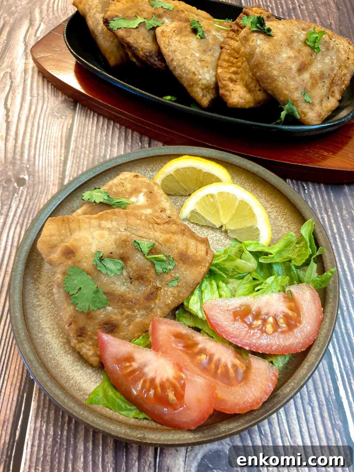 2 samosas on a brown plate with lettuce, slices of tomato  and lemon wedges.
