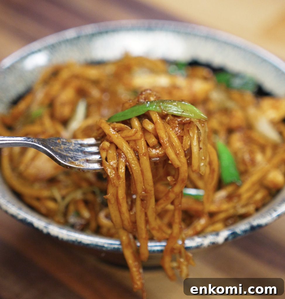 Crispy Chicken Pan-Fried Noodles with Garlic Scallions 3 A fork lifts a glossy bite of pan fried noodles with chicken and scallions from the bowl. The noodles are coated in a rich, savory sauce that clings to every strand.