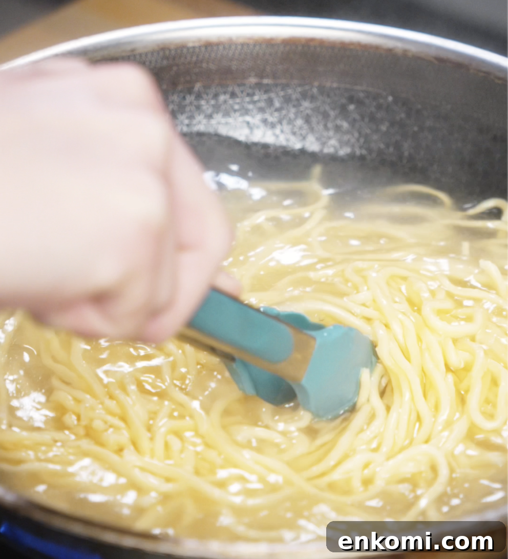 Crispy Chicken Pan-Fried Noodles with Garlic Scallions 5 Fresh wheat noodles are boiling in a pot of water, stirred with tongs to prevent sticking. These chewy noodles form the base of the pan-fried dish.