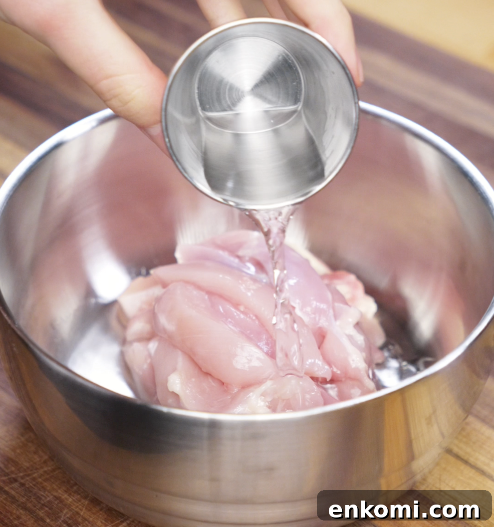 Crispy Chicken Pan-Fried Noodles with Garlic Scallions 6 A hand pours Shaoxing wine over raw sliced chicken thighs in a metal bowl. This step helps tenderize the meat and infuse it with classic Chinese flavor.