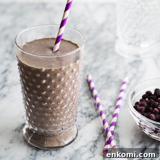High-Protein Chocolate Blueberry Smoothie Recipe 2 A chocolate blueberry smoothie in a glass with purple striped straws and blueberries beside.
