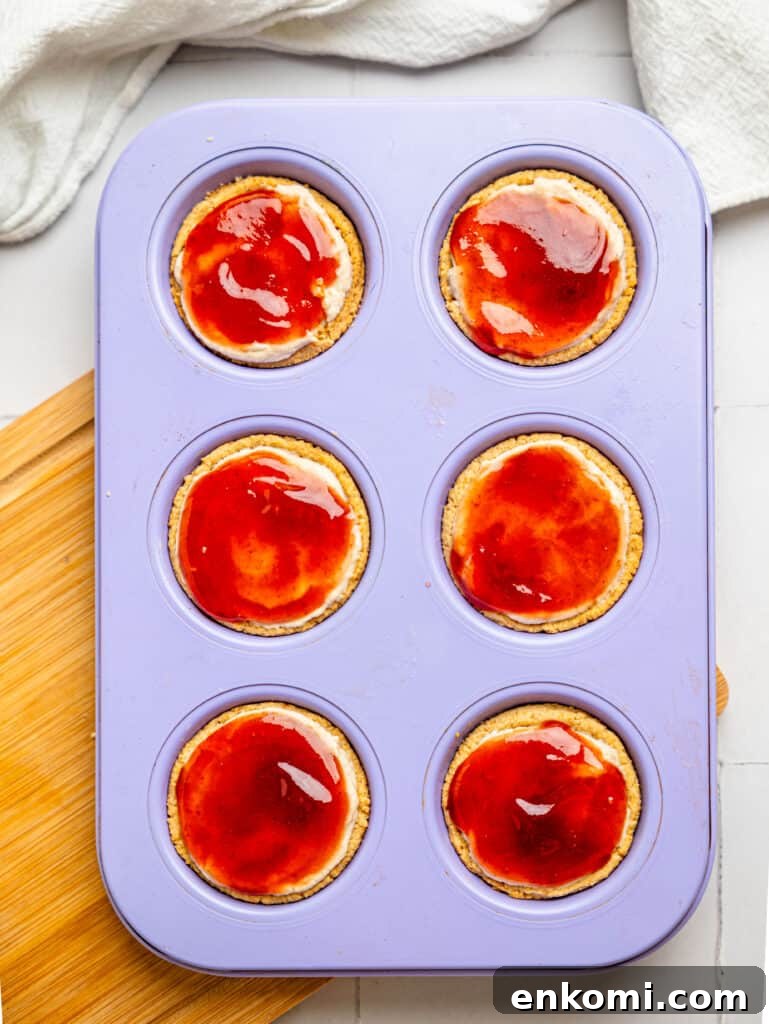 High-Protein Mini Cheesecakes: Low-Calorie Dessert Recipes 9 Mini cheesecake crusts filled with protein cheesecake filling with strawberry jam on top in a muffin tray.