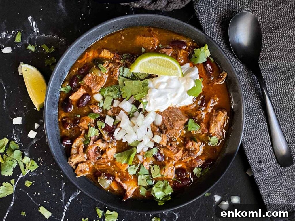 Smoked pork chili garnished with cilantro, onion, lime, sour cream, and onion in a dark bowl.