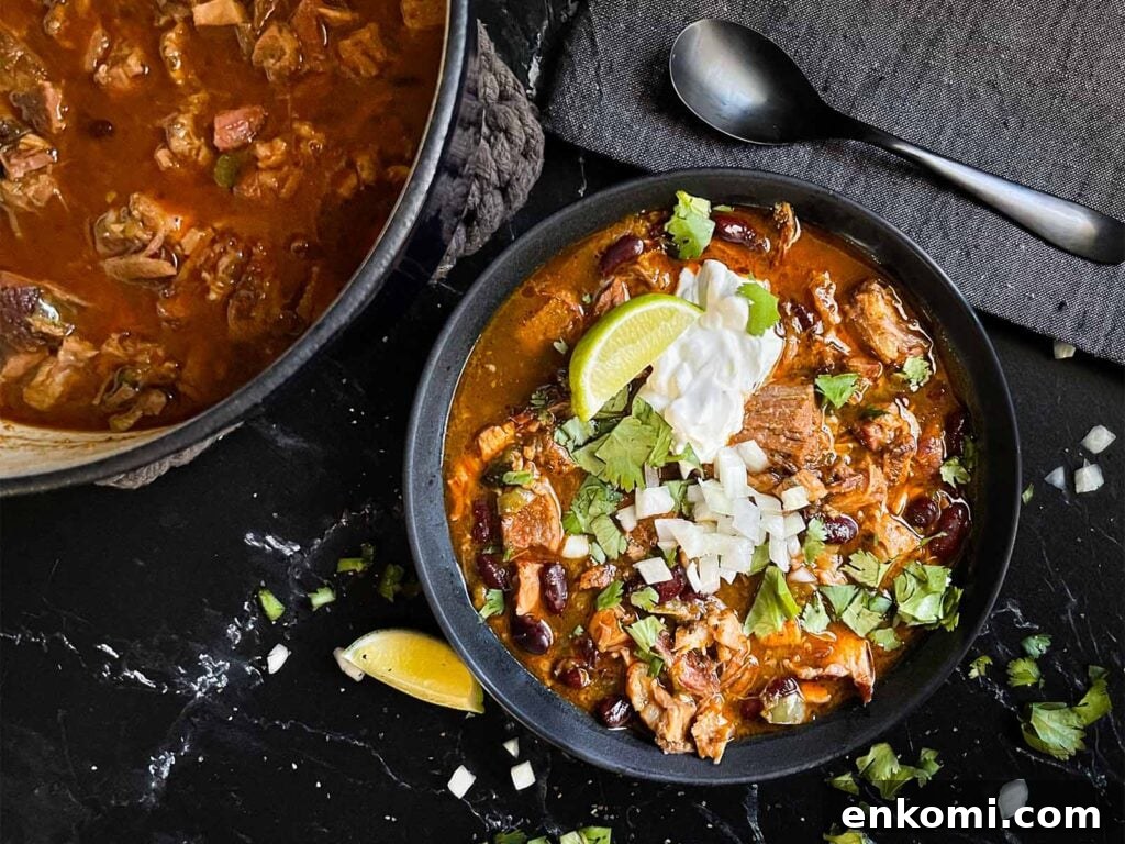 Smoked pork chili garnished with cilantro, onion, lime, sour cream, and onion in a dark bowl.