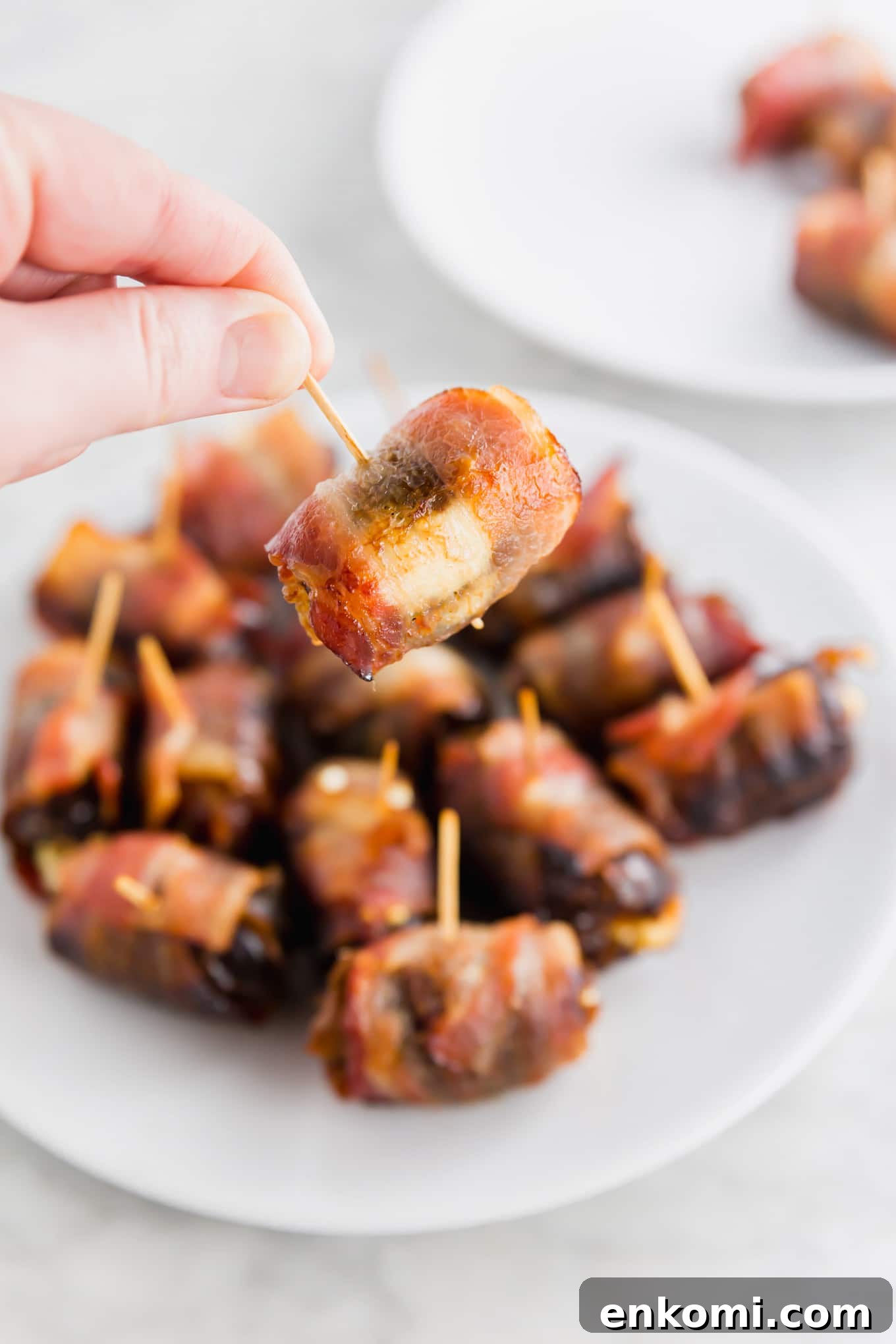 Bacon-Wrapped Dates Stuffed with Goat Cheese and Honey 5 A photo of a hand holding a bacon wrapped date over a platter of goat cheese stuffed dates.