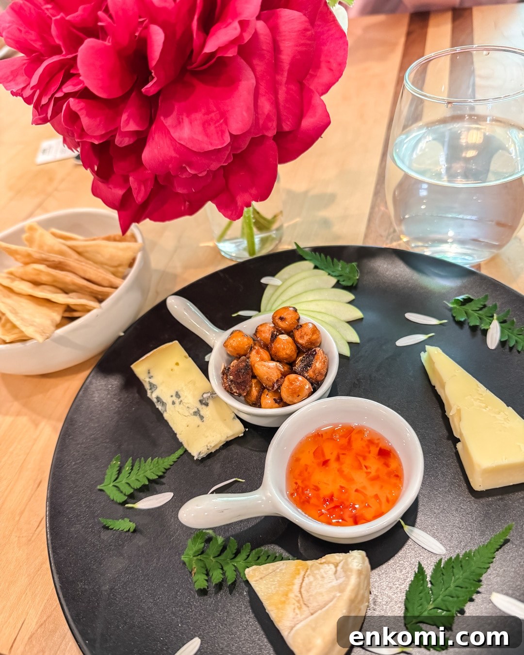 A black plate with cheese wedges, sweet chili sauce, roasted chickpeas, apple slices, and ferns. Crackers in a bowl, a pink peony, and a glass of water in the background.