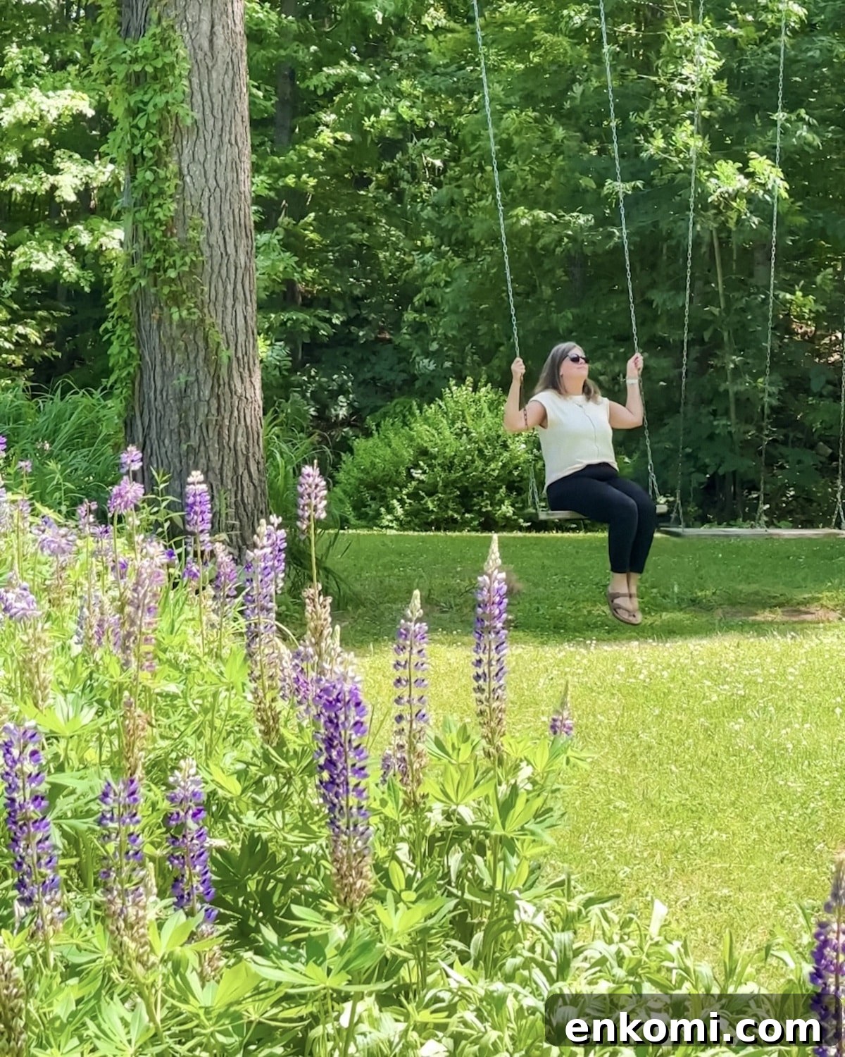 A woman sits on a swing hanging from a tree in a sunny garden with purple lupine flowers in the foreground.