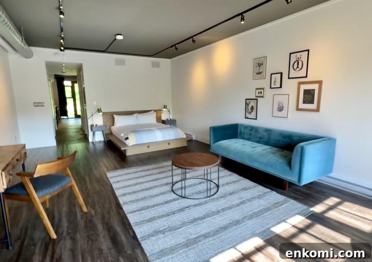 A modern bedroom features a bed, teal sofa, round coffee table, desk with chair, and wall art, with sunlight streaming onto a striped rug over wood floors.