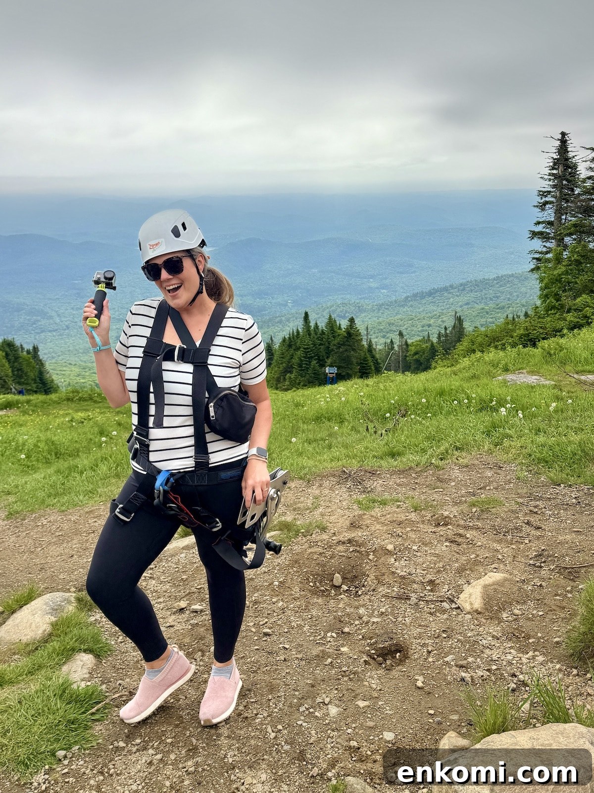 A woman wearing a helmet and harness stands on a mountain trail holding a small camera, with green hills and cloudy sky in the background.