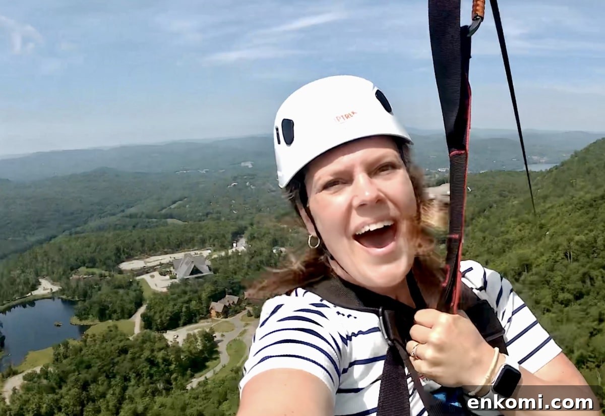 A woman wearing a helmet and striped shirt smiles while ziplining high above a forested landscape with hills, a lake, and buildings visible below.