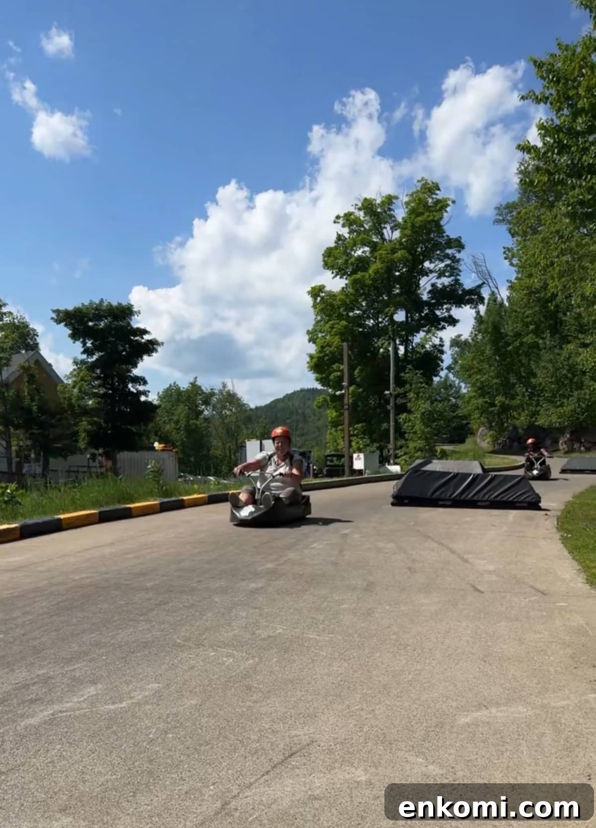 A person wearing a helmet rides a luge cart down a paved outdoor track on a sunny day, with trees and hills in the background.