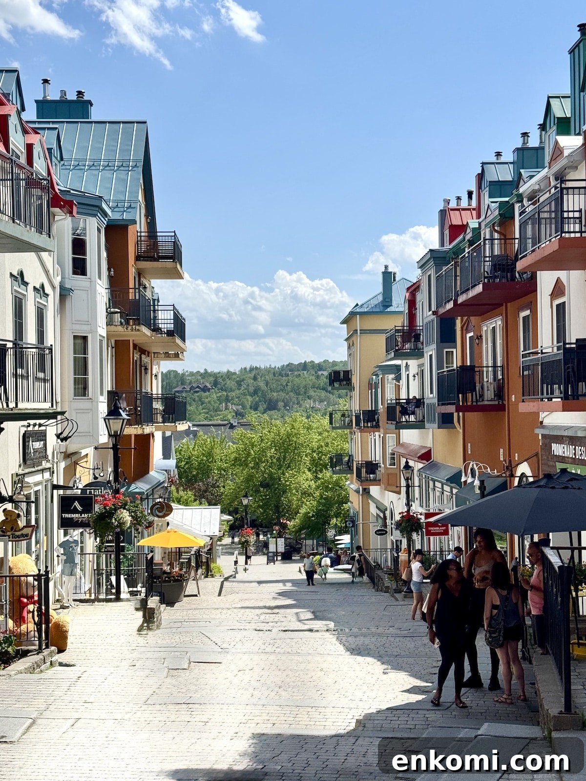 A cobblestone street lined with colorful buildings and balconies on both sides, with people walking and green hills visible in the background under a blue sky.