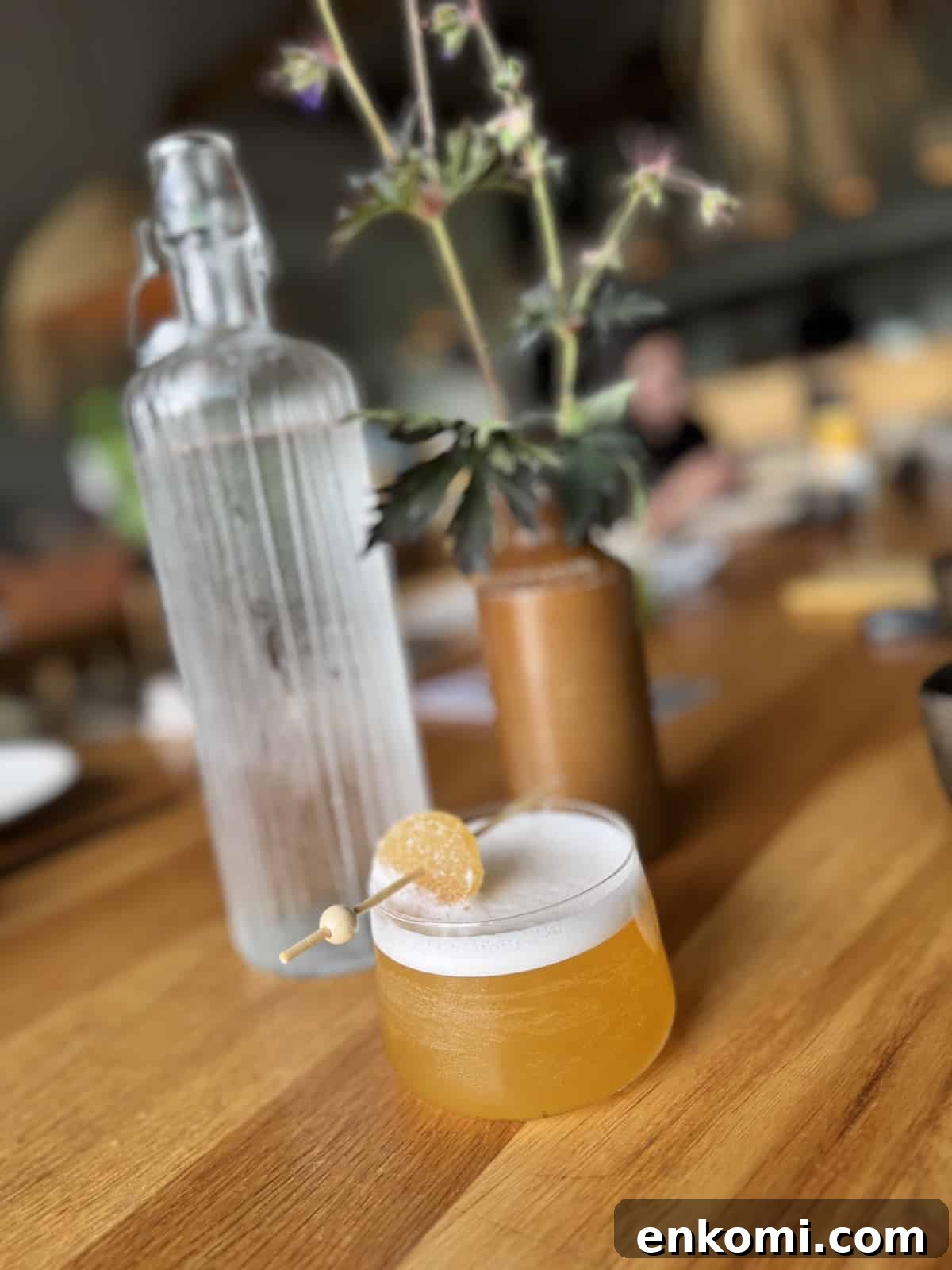 A frothy yellow cocktail garnished with a candied twist sits on a wooden table next to a tall glass water bottle and a brown vase with flowers.