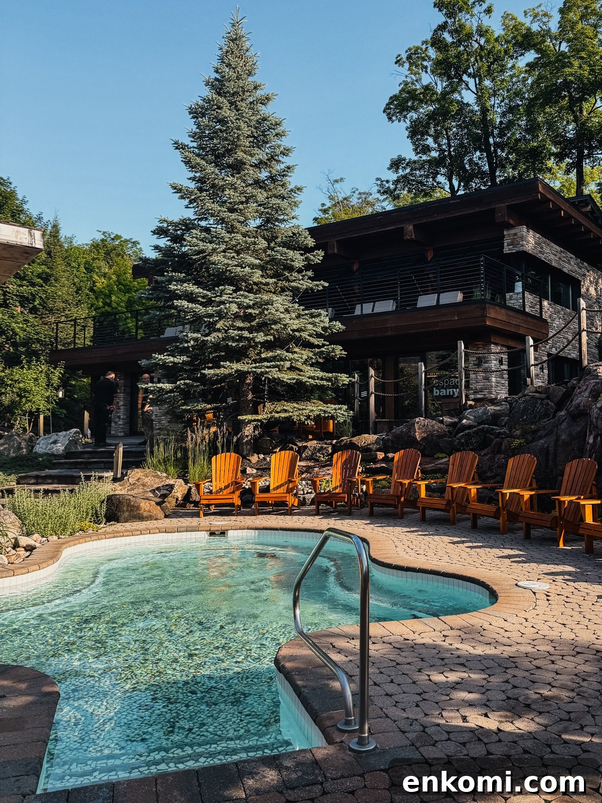 A small outdoor pool with clear water, surrounded by orange wooden chairs and stone paving, sits in front of a modern house and tall pine tree, under a sunny sky.
