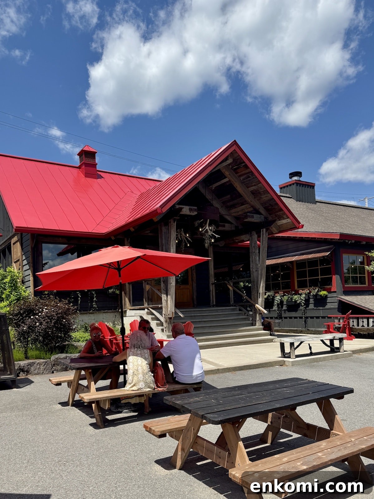 Three people sit at a wooden picnic table under a red umbrella outside a rustic building with a bright red roof on a sunny day.