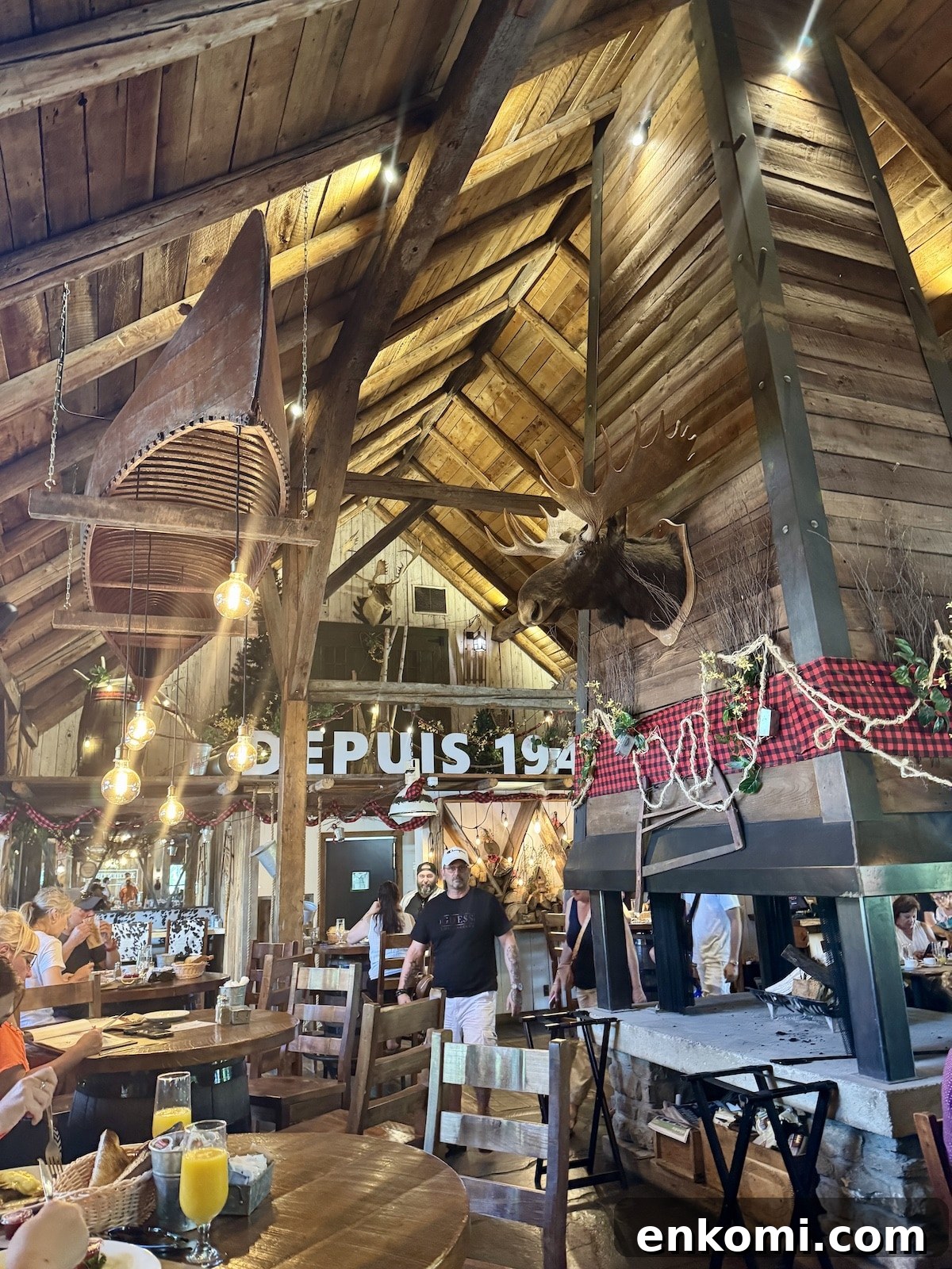 Interior of a rustic restaurant with wooden beams, mounted moose head, hanging lights, and diners seated at tables. A sign on the back wall reads “DEPUIS 194.”.