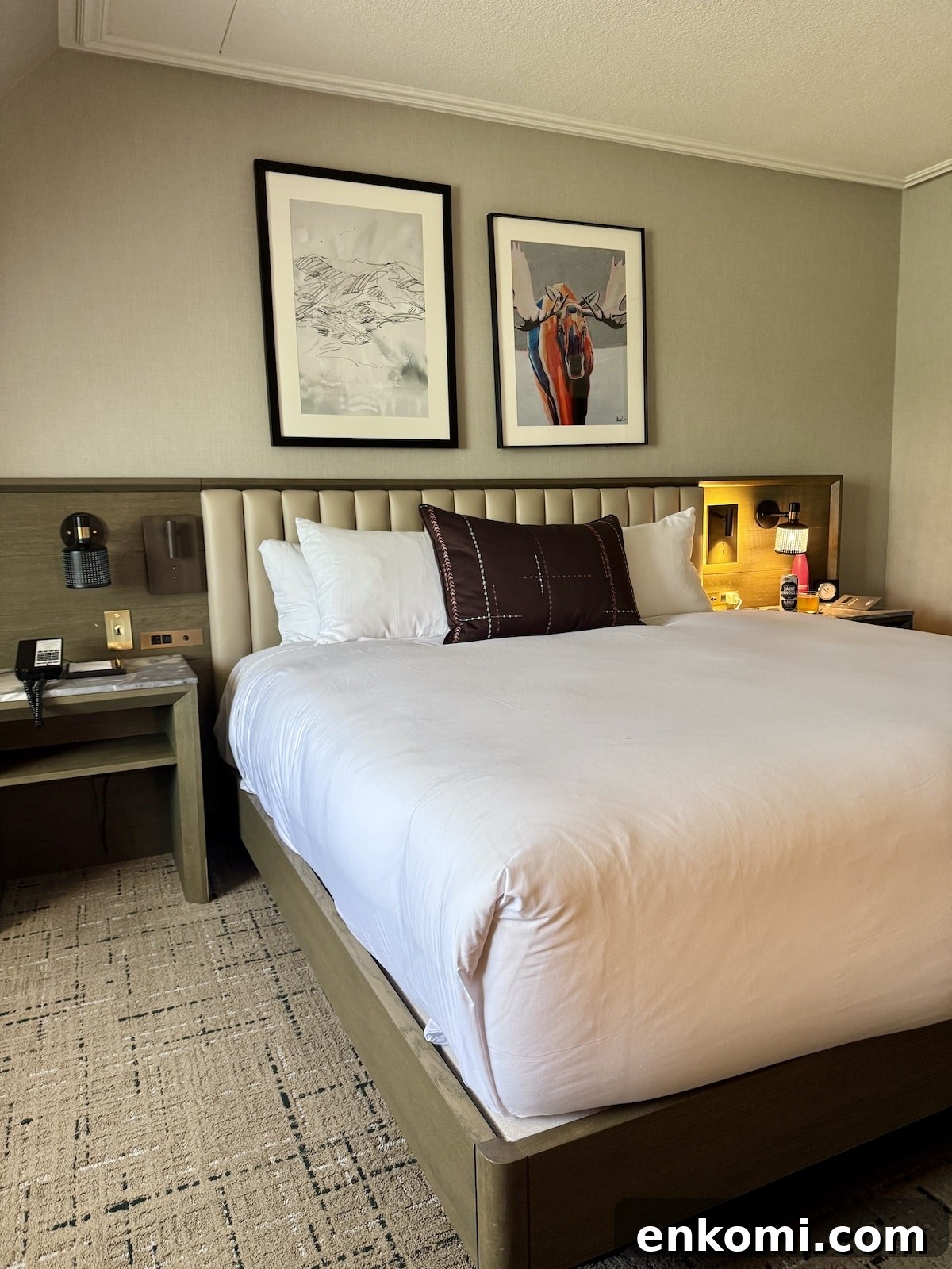 A neatly made hotel bed with white linens, two framed artworks above the headboard, and a nightstand with a lamp, phone, and water bottle.