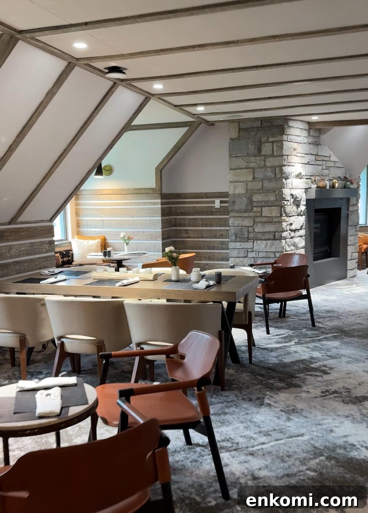 Modern dining room with beige chairs, set tables, a stone fireplace, and angled ceiling. Soft lighting and minimalist decor create a cozy, contemporary atmosphere.