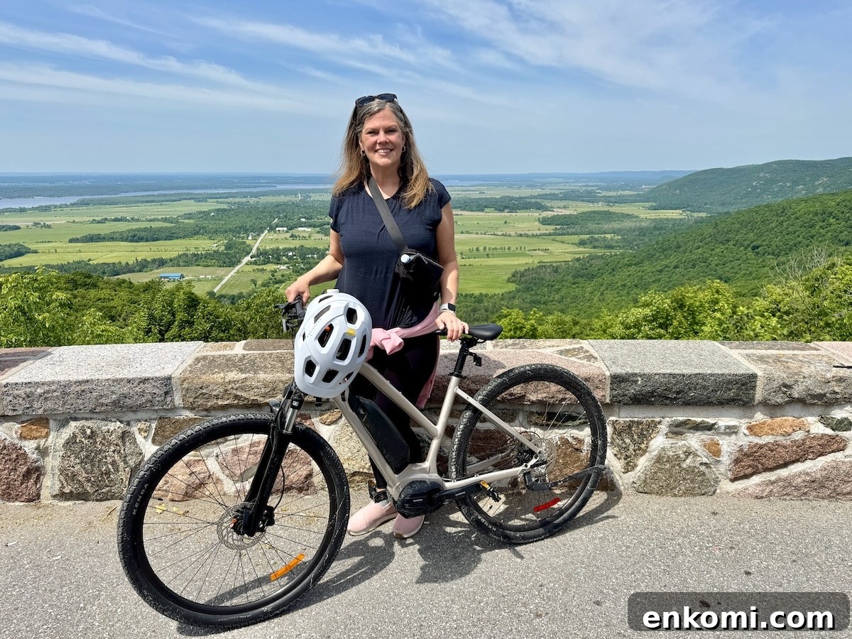 A woman stands behind a bicycle with a white helmet resting on the handlebars, posing in front of a stone wall overlooking a scenic valley and rolling hills under a blue sky.
