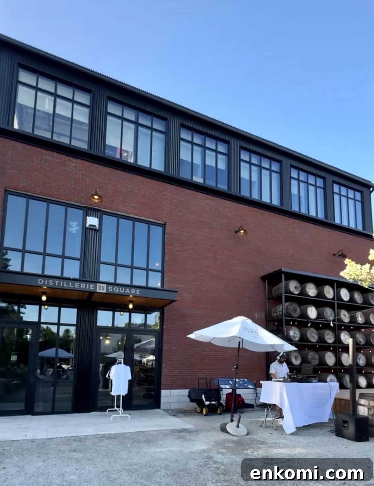 A red brick distillery building with large windows, stacked barrels on the side, and a white tented table set up outside on a sunny day.